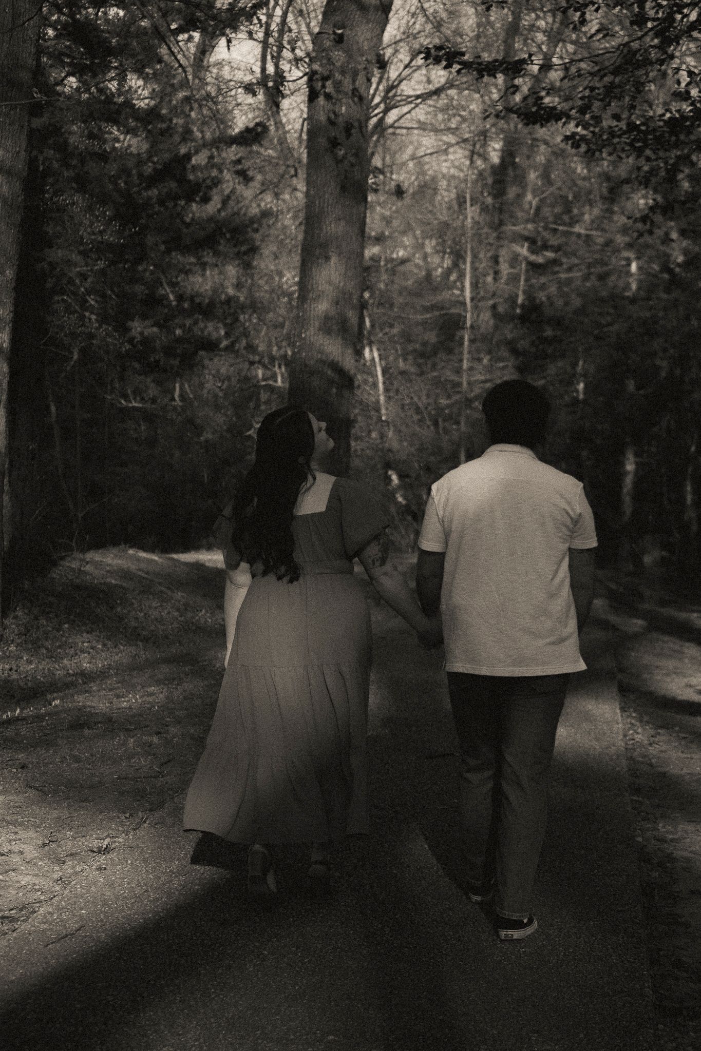 Black and white photo of a couple walking hand in hand down a forest path surrounded by tall trees.