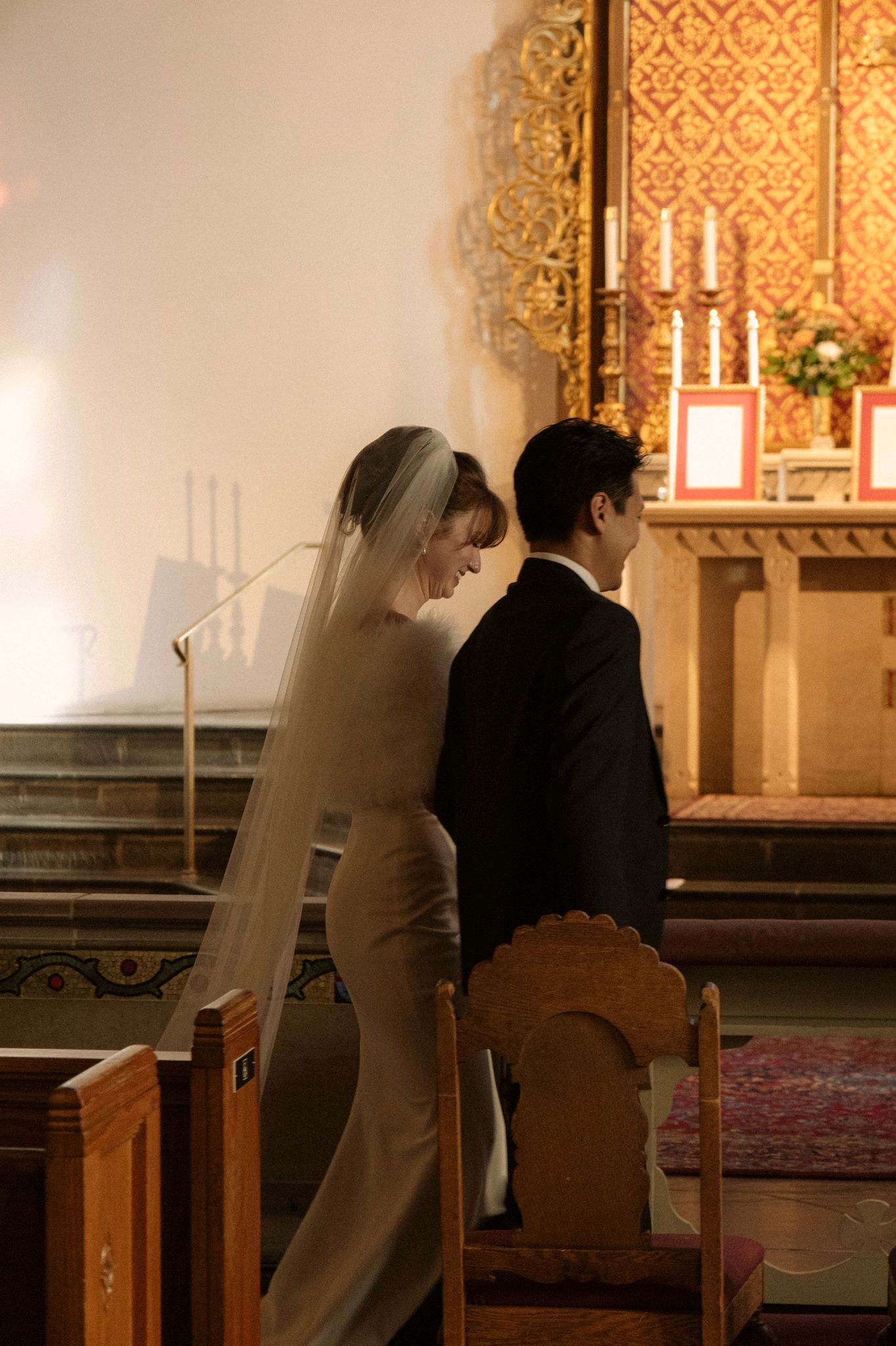 Bride in long veil and groom in dark suit kneel together before ornate church altar during ceremony.