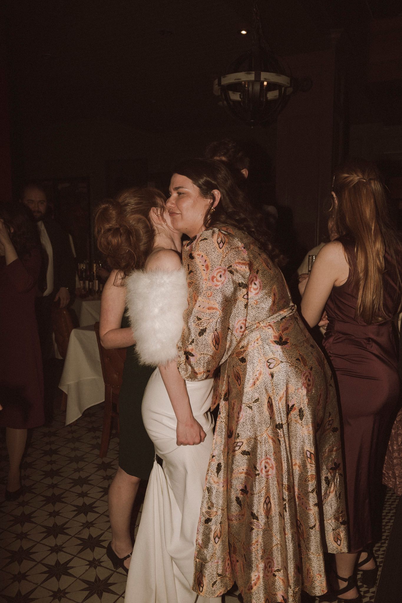 Bride in white gown with feather wrap receives a kiss on the cheek from a guest at reception.