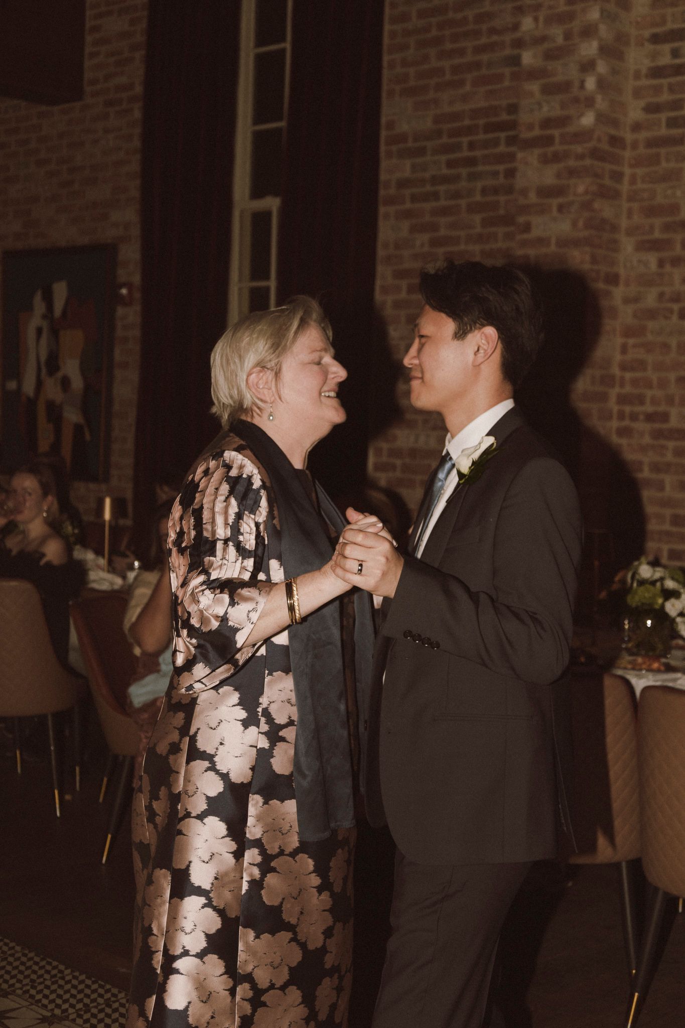 A groom in a dark suit shares a joyful mother-son dance with a woman in a floral gown at a wedding reception.