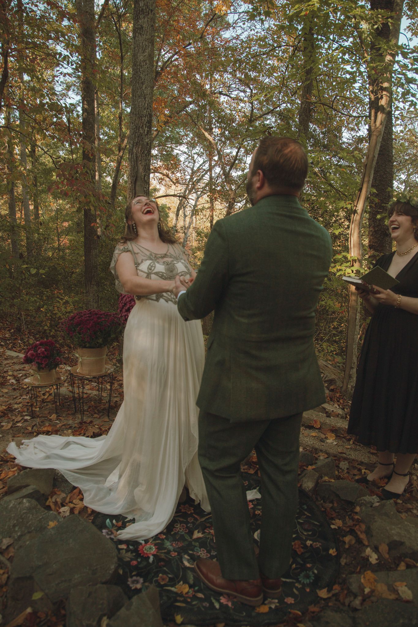 A couple shares a joyful moment during their autumn woodland wedding ceremony with fallen leaves on the ground.