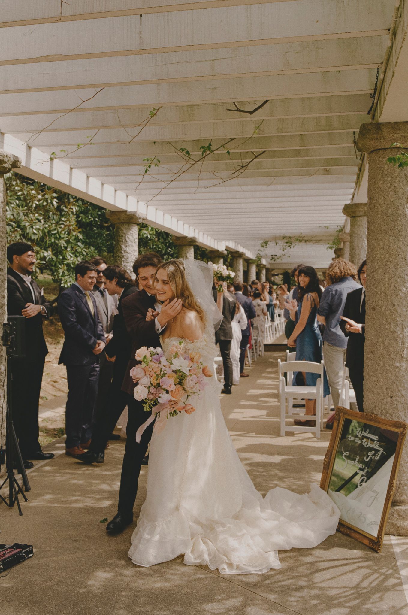 Wedding guests gather in a covered outdoor colonnade area with white columns and ceiling during a celebration.