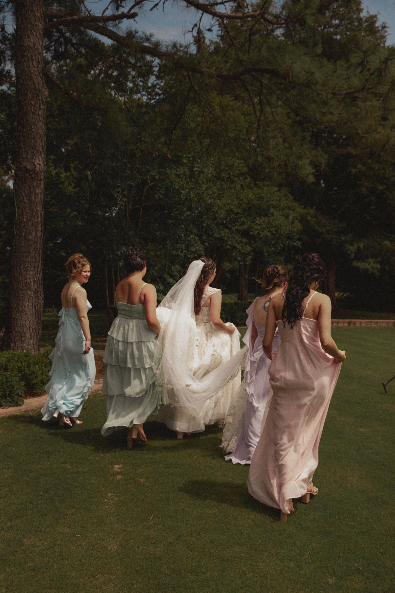 A group of women in flowing dresses run across a grassy lawn in a whimsical outdoor photo shoot.