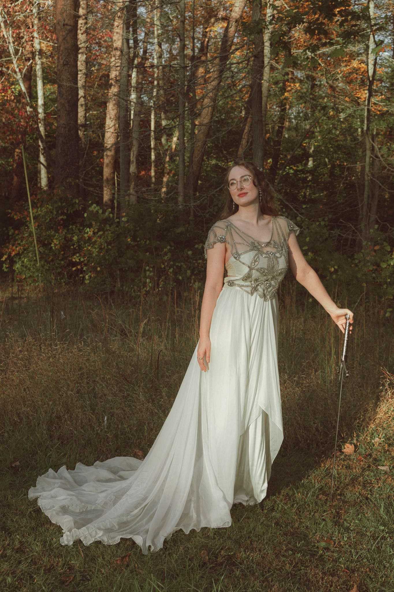 Ethereal figure in flowing white dress poses gracefully among autumn trees at golden hour in woodland setting.