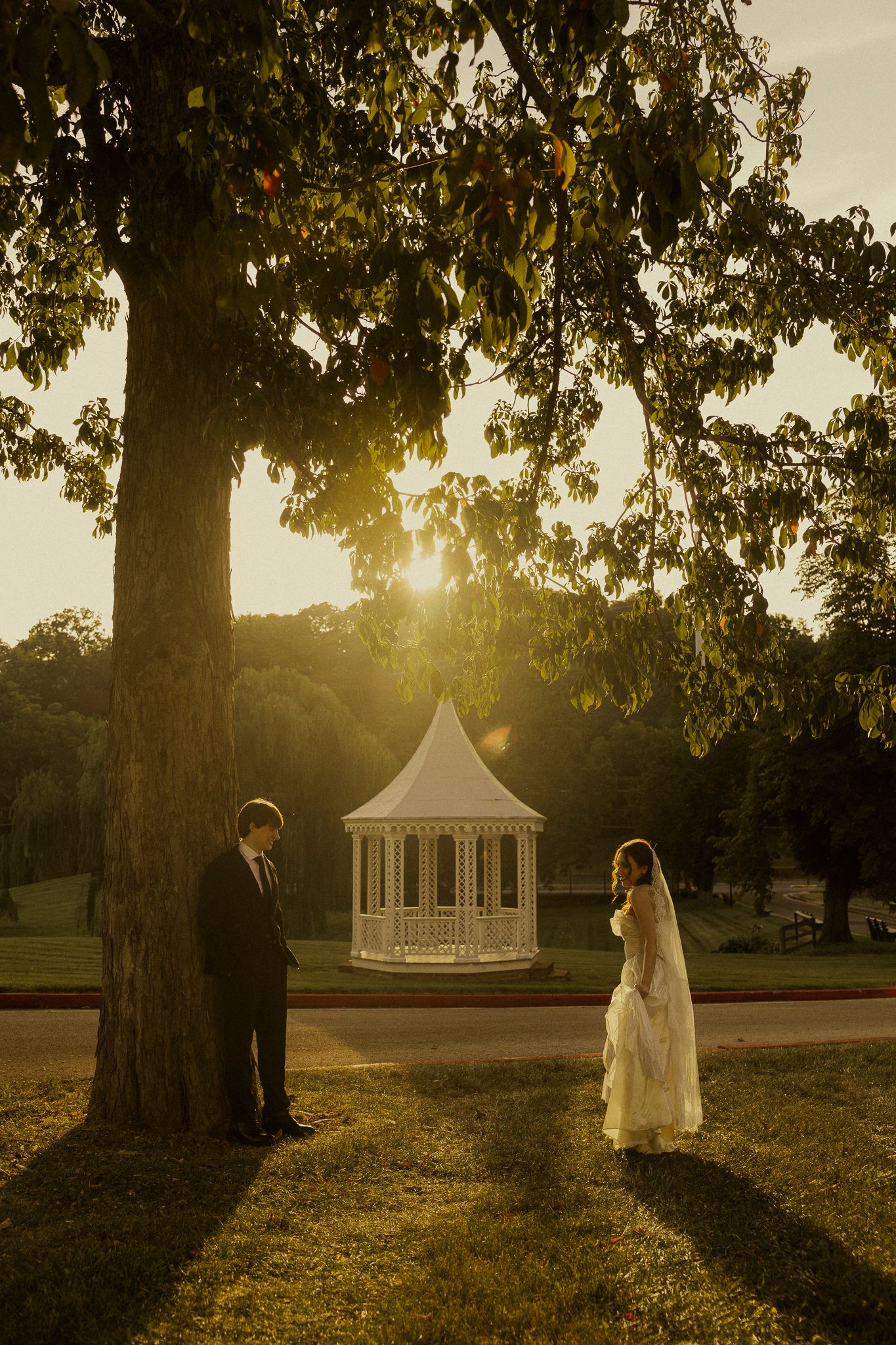 Bride and groom share a romantic golden hour moment under a large tree near a white gazebo in a lush park.