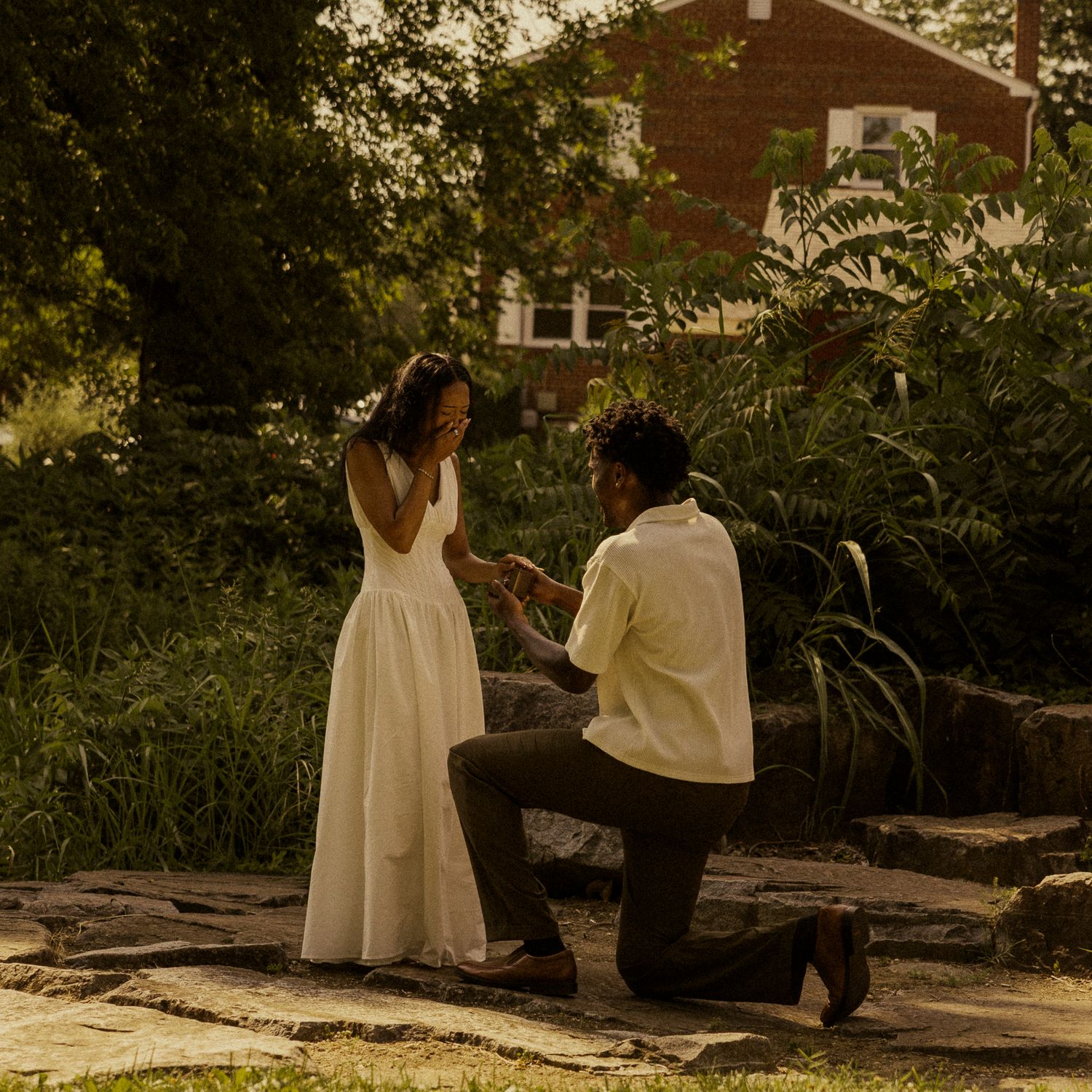 Man kneeling on one knee proposing to woman in white dress in a lush garden near a brick building.