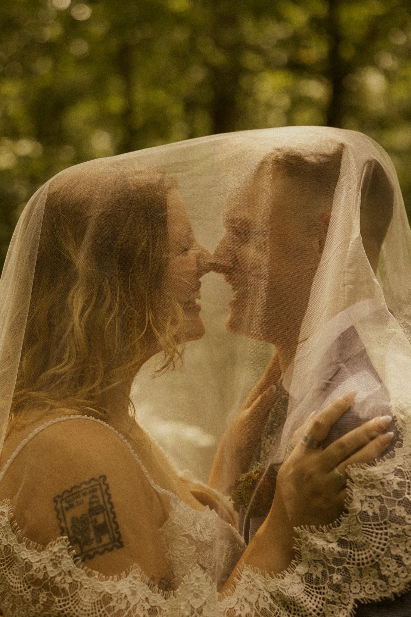 Bride and groom sharing a romantic moment under a wedding veil in a lush green forest setting.