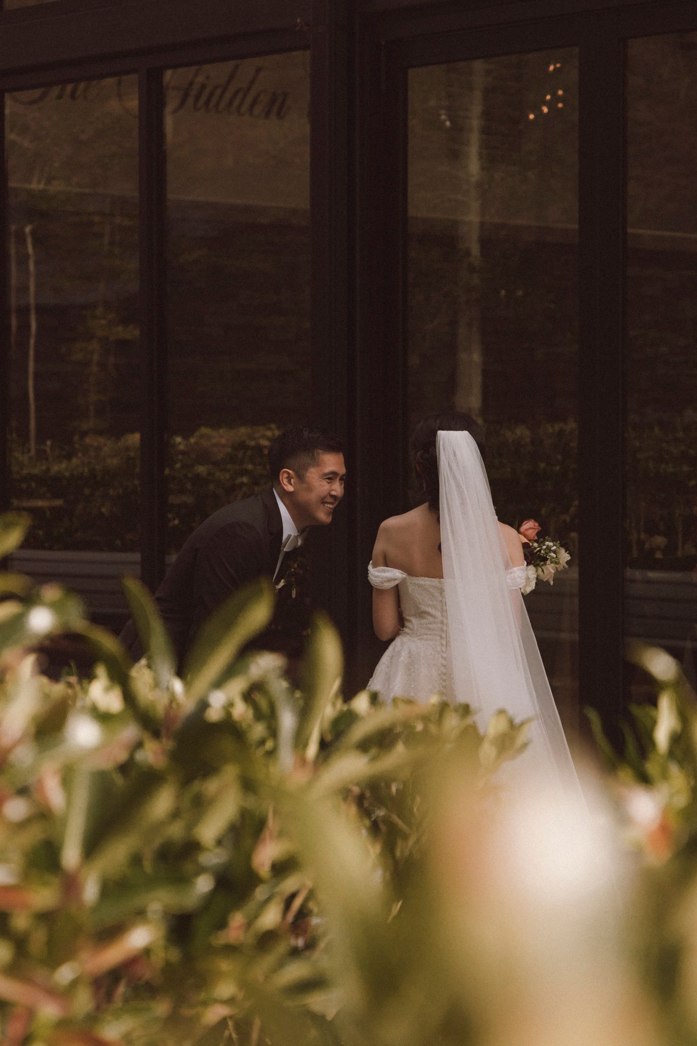 A bride and groom share an intimate moment in a garden setting with soft bokeh lighting and greenery in the foreground.
