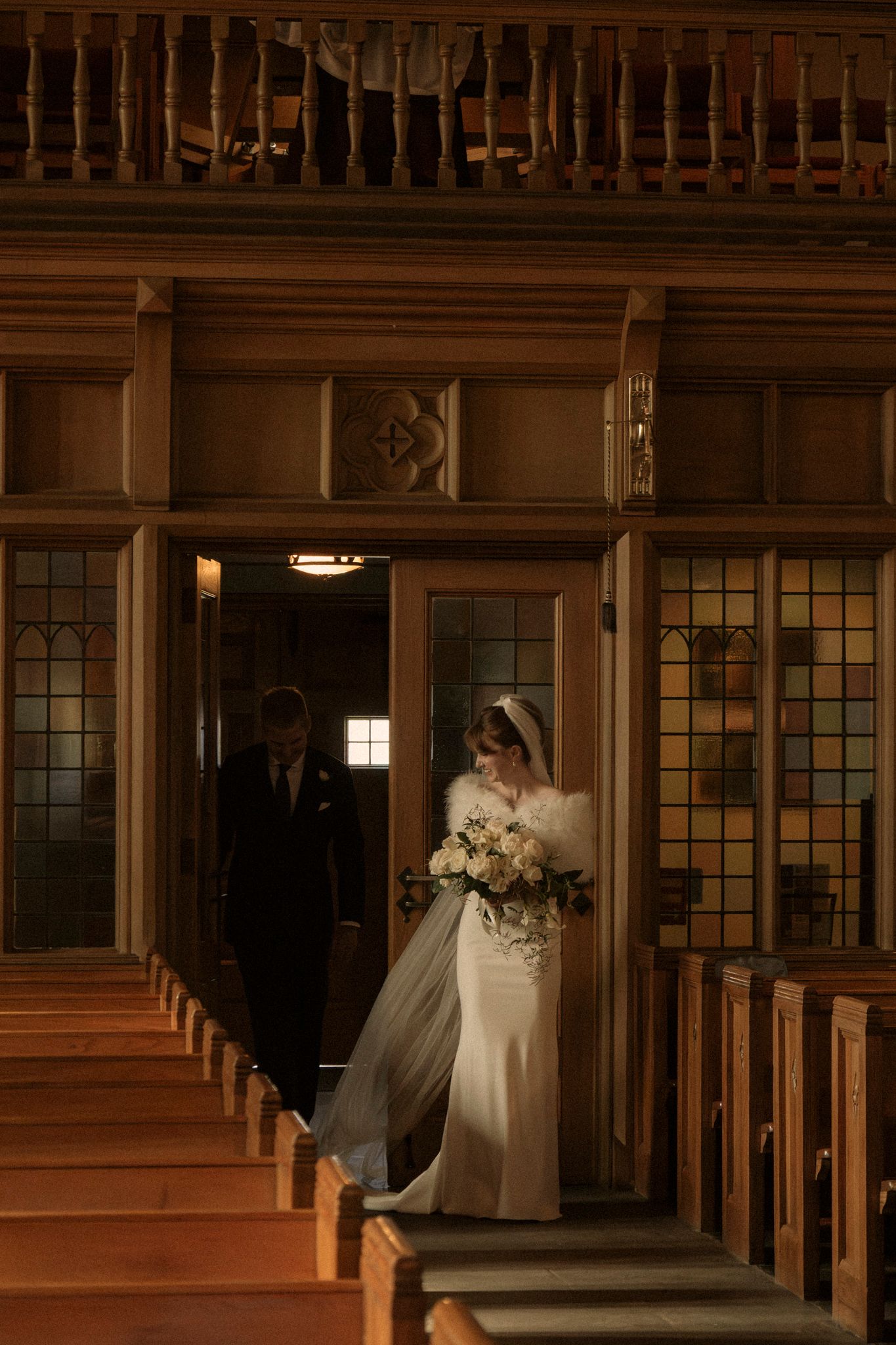 A couple stands in a warmly lit wooden church doorway during a wedding ceremony, with pews visible in the foreground.