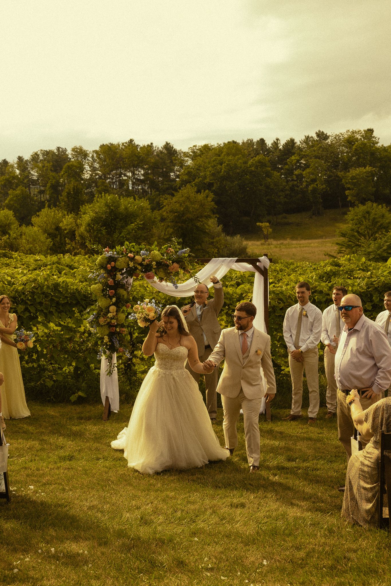 Bride and groom walk hand-in-hand down the aisle at an outdoor vineyard wedding ceremony.