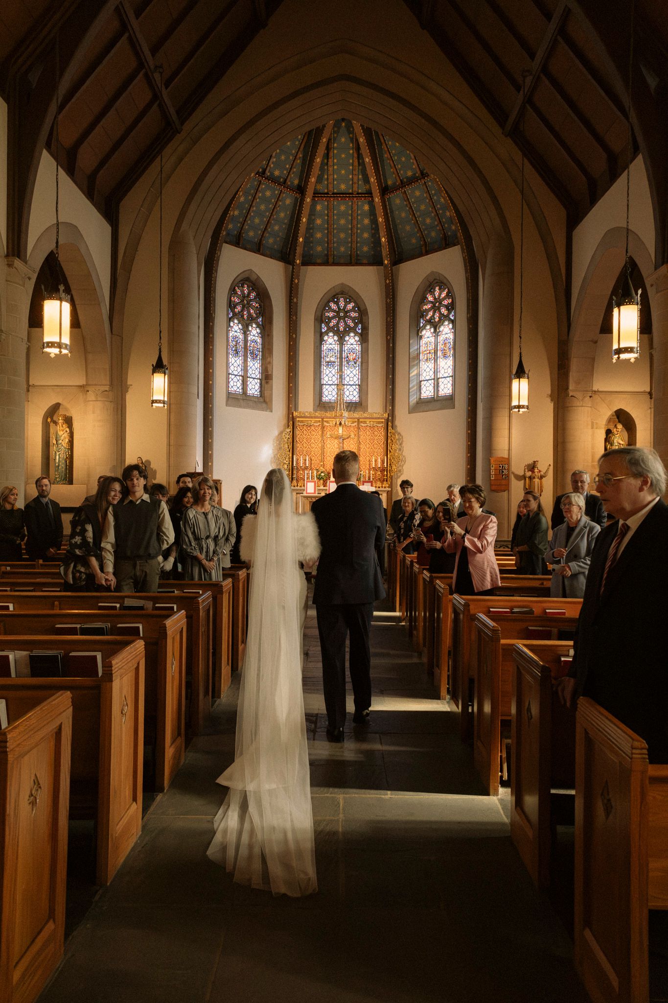 Bride walking down the aisle with escort in a beautiful church wedding ceremony with stained glass windows.
