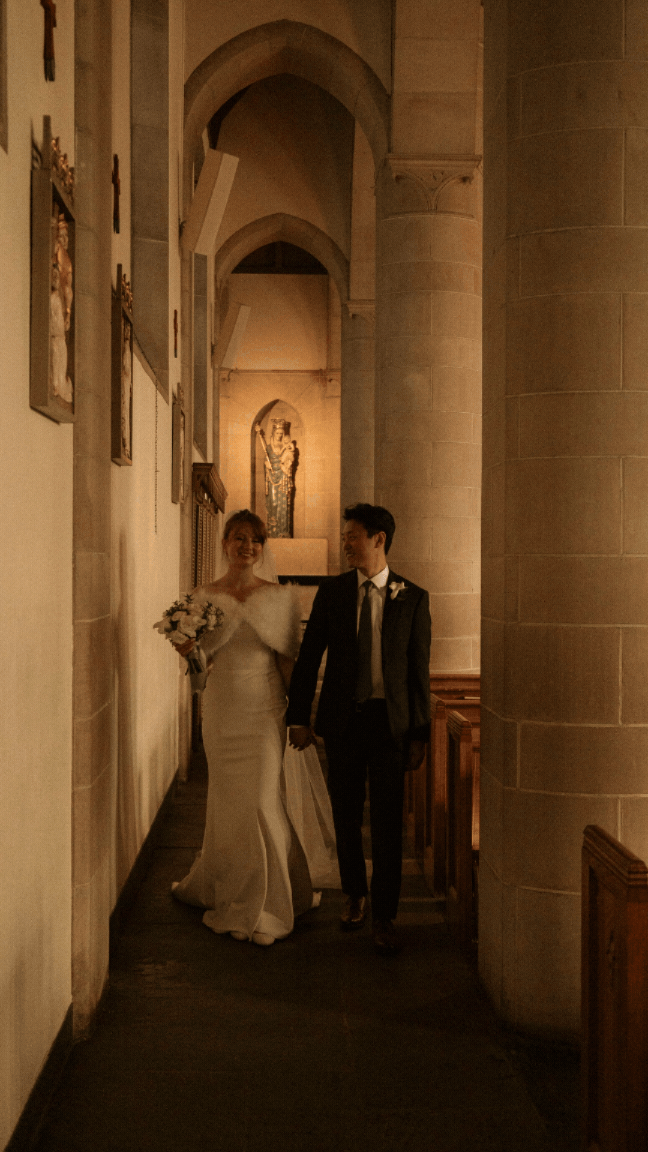 A bride and groom walk hand in hand through a warmly lit stone church corridor, the bride in a white gown holding a bouquet.