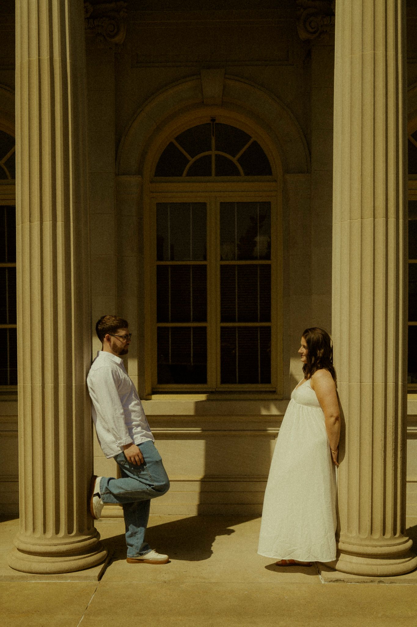 Pregnant woman in white dress and man in white shirt stand between stone columns of an elegant neoclassical building.