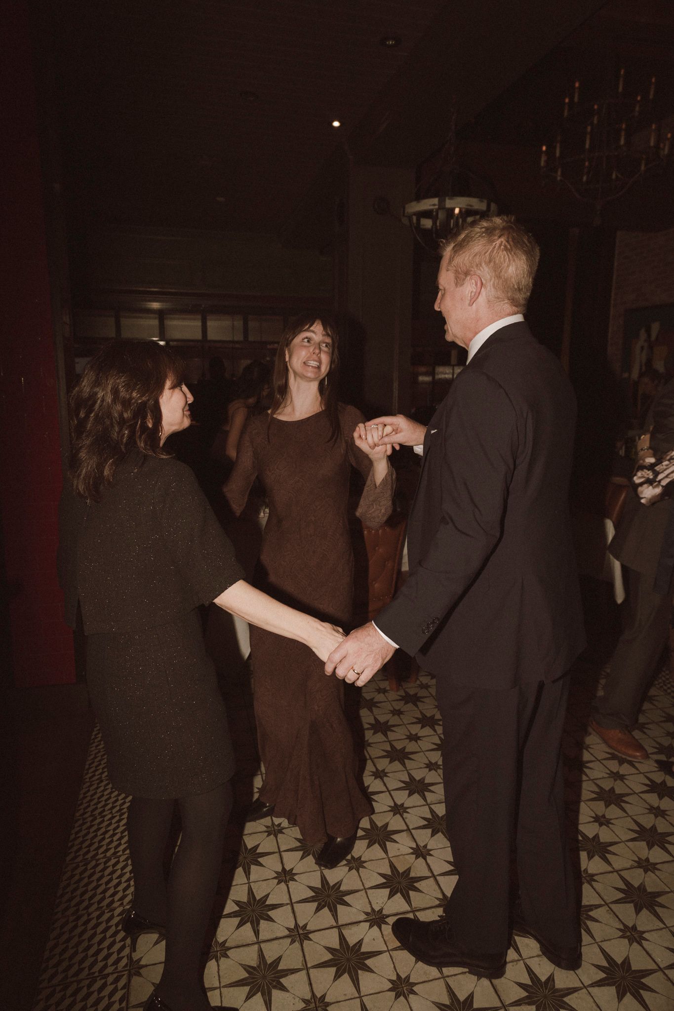 Guests in formal attire hold hands and socialize on a patterned tile floor at a wedding reception.