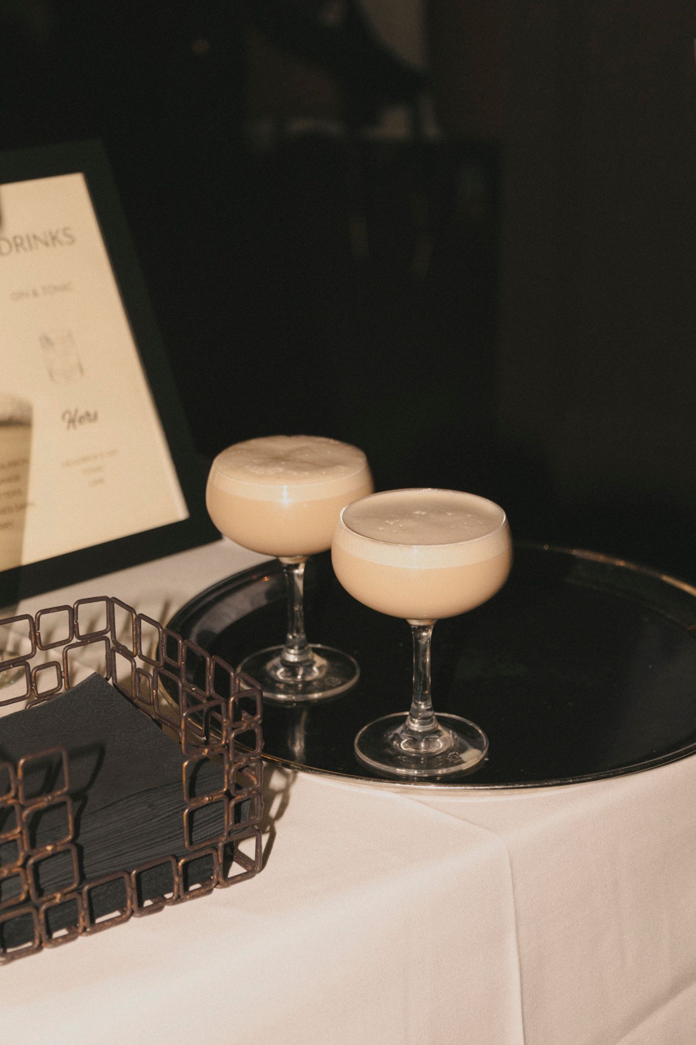 Two creamy foam-topped cocktails served in coupe glasses on a black tray at a wedding reception bar.