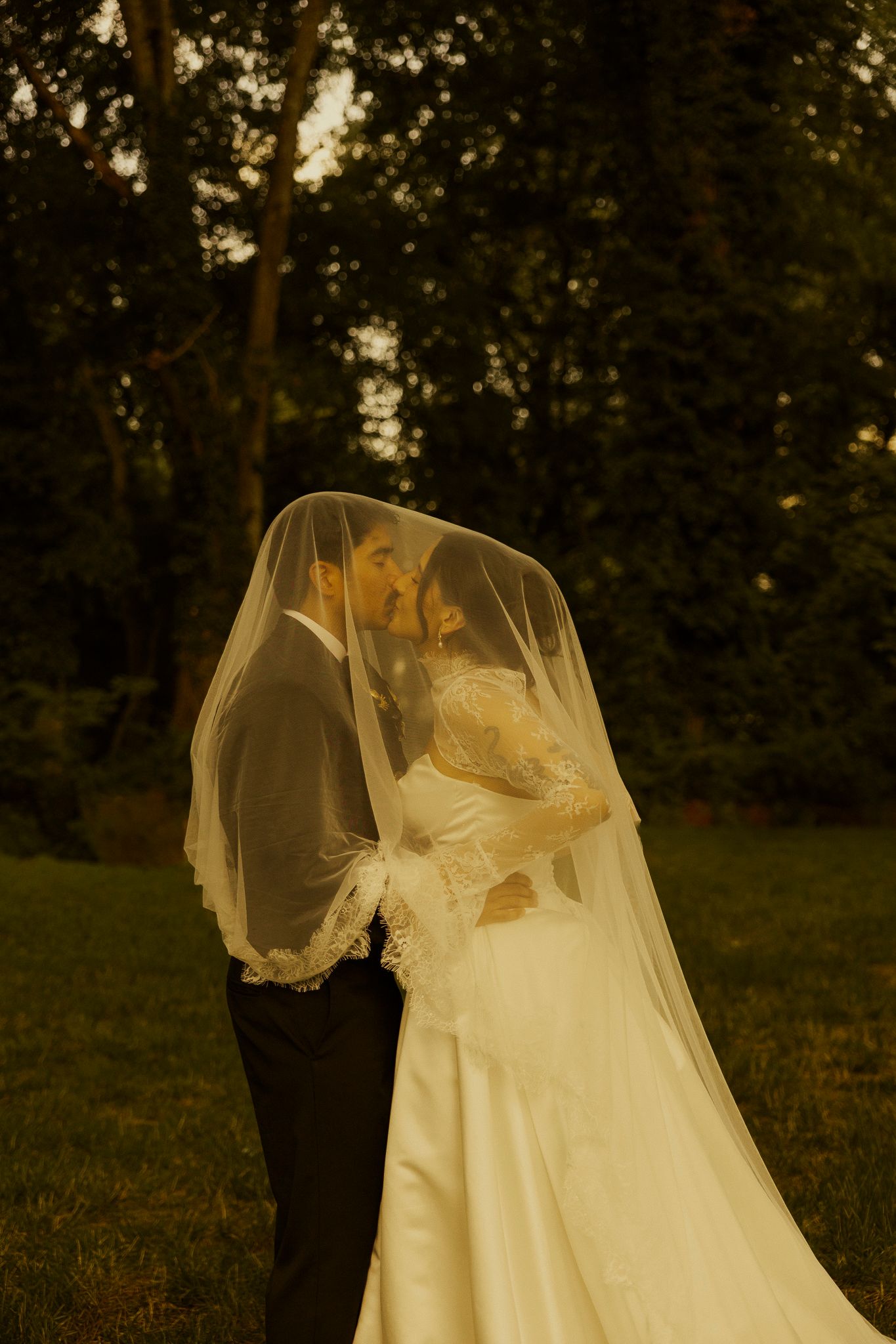 Bride and groom sharing a kiss under a flowing veil in a lush green garden at golden hour.