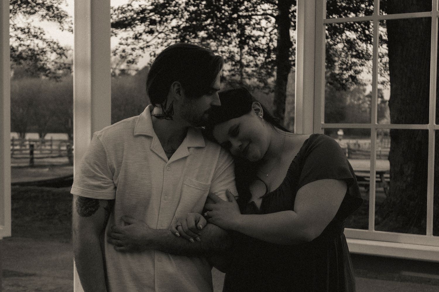 Black and white photo of a romantic couple standing close together near elegant white French doors outdoors.