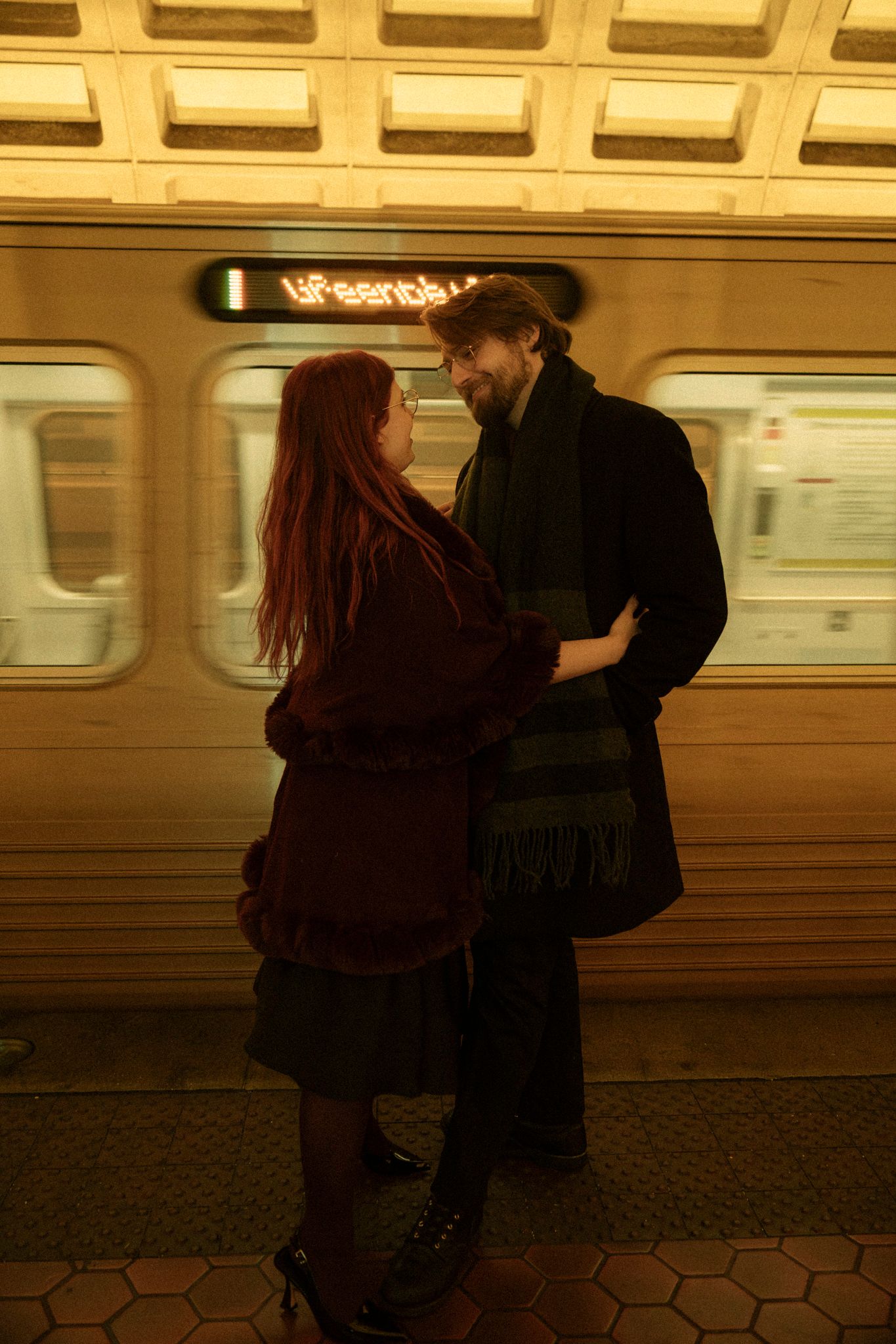 Two people embrace on a subway platform with a train car visible in the warm-toned background.