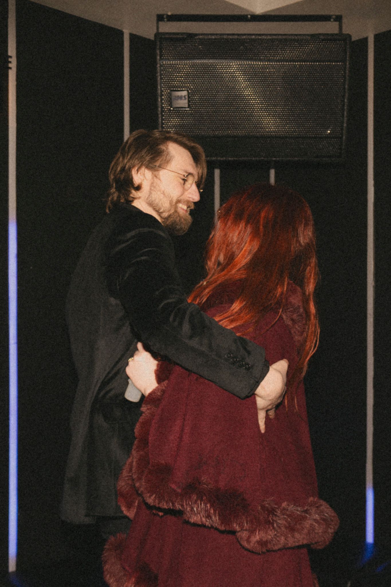 A couple embraces in a dimly lit room, with a speaker visible on the wall behind them.