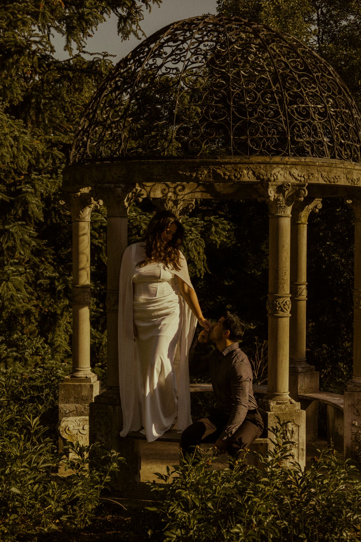 Couple in romantic garden gazebo, woman in white dress standing as man kneels beside her, surrounded by lush greenery.
