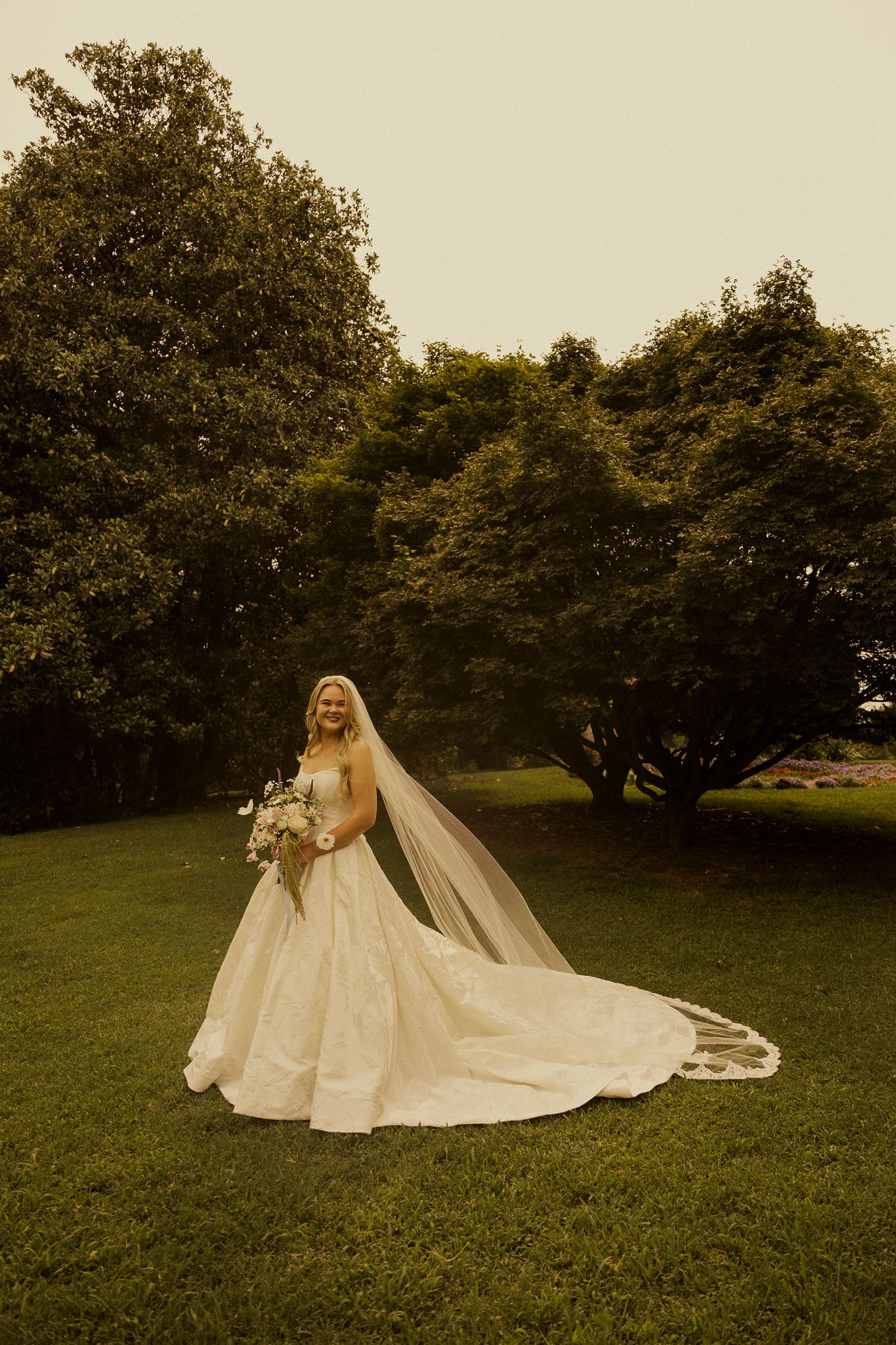 Bride in a flowing white ball gown and long veil holding a bouquet, standing on green grass with lush trees behind her.