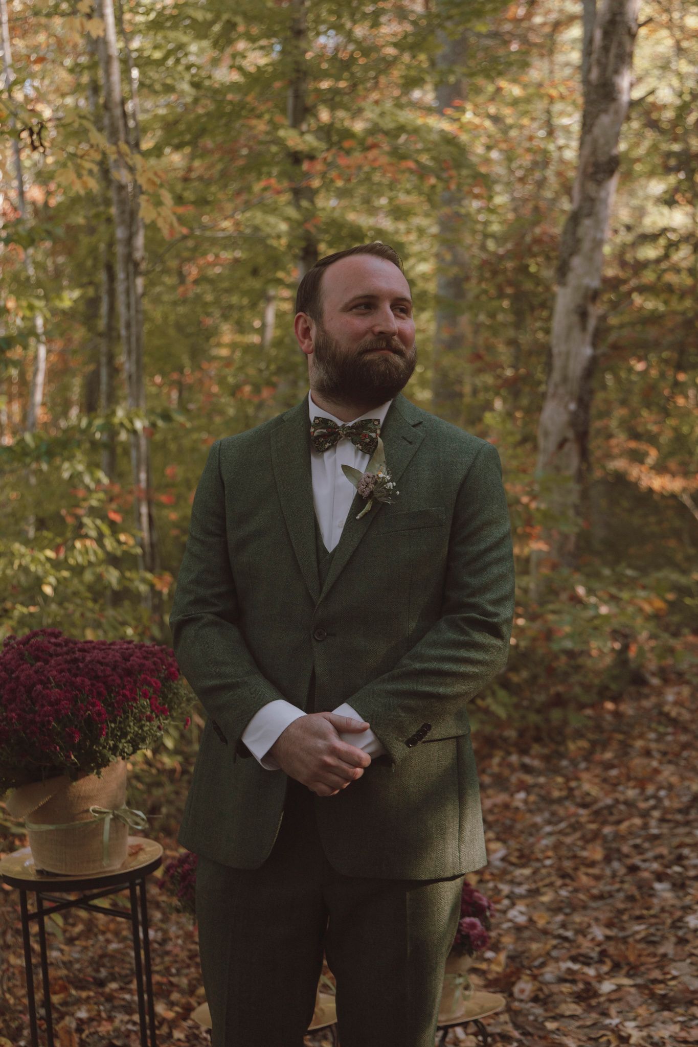 Person in olive green suit stands in autumn forest setting with fallen leaves and muted natural lighting.