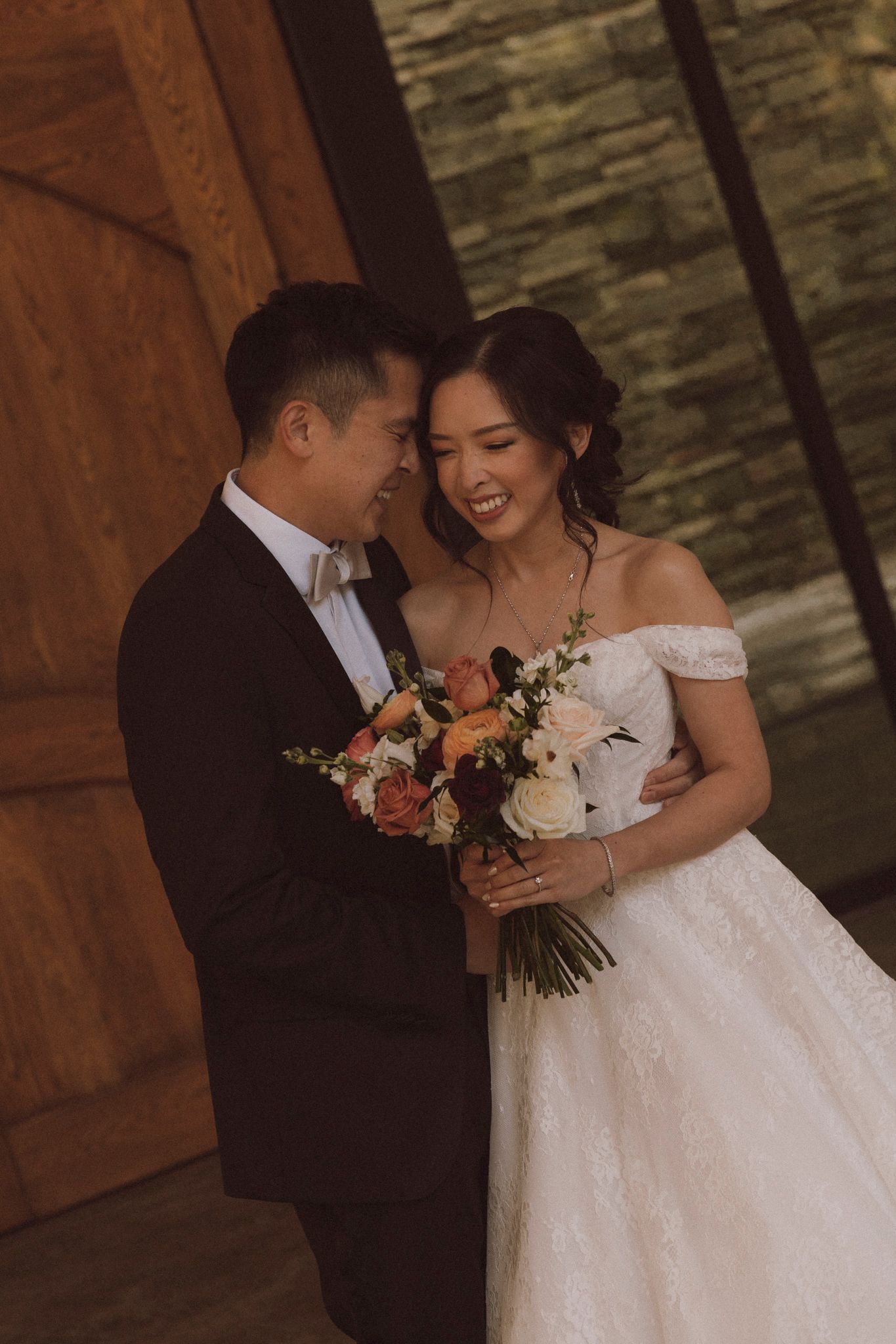 A romantic wedding moment captured as newlyweds share a tender moment with a floral bouquet against a rustic stone wall.