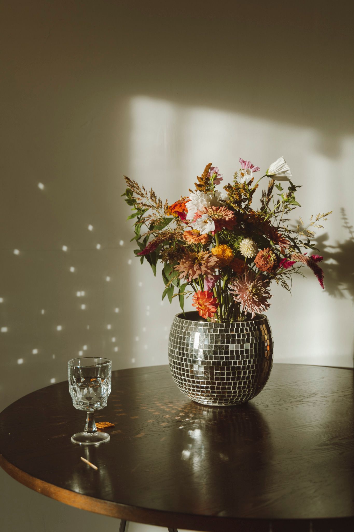 Disco ball vase with autumn flowers creates sparkly light patterns on wall while sitting on dark wooden table with crystal glass.