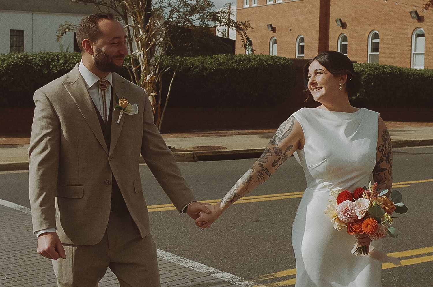 Joyful wedding couple holding hands while walking down a sunny street with brick buildings.