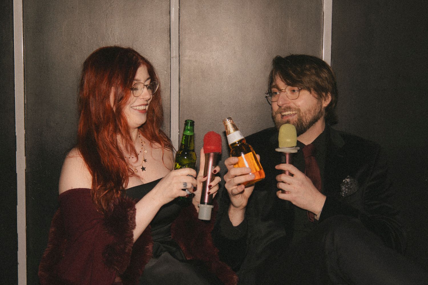 Two people share drinks and conversation in a dimly lit setting with dark background.