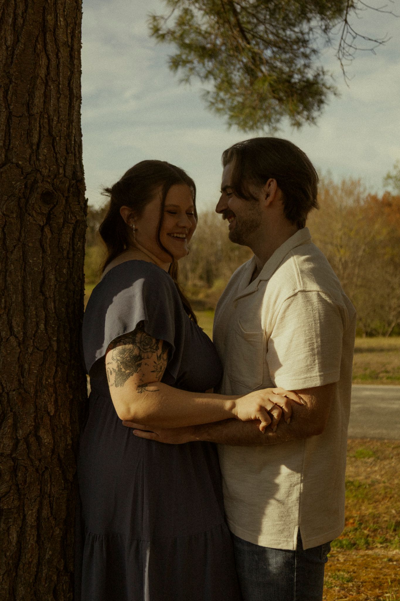 A smiling couple embraces near a tree in golden sunlight, the woman wearing a gray dress with a tattoo visible on her arm.