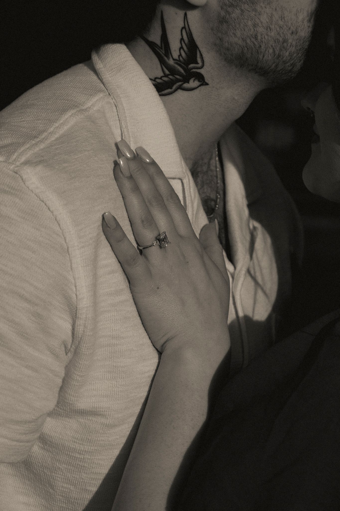 Close-up black and white photo of a hand with a bow ring resting on a mans chest with a bird tattoo.