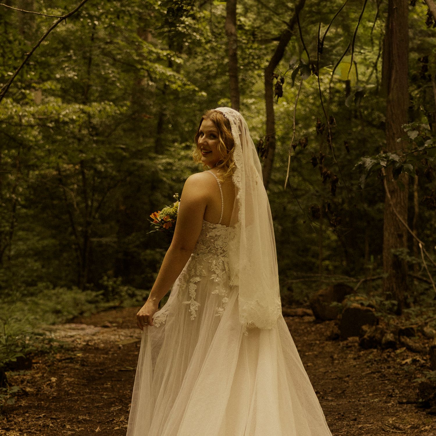 A bride in a white gown and veil walks through a lush forest path, holding a bouquet of flowers.