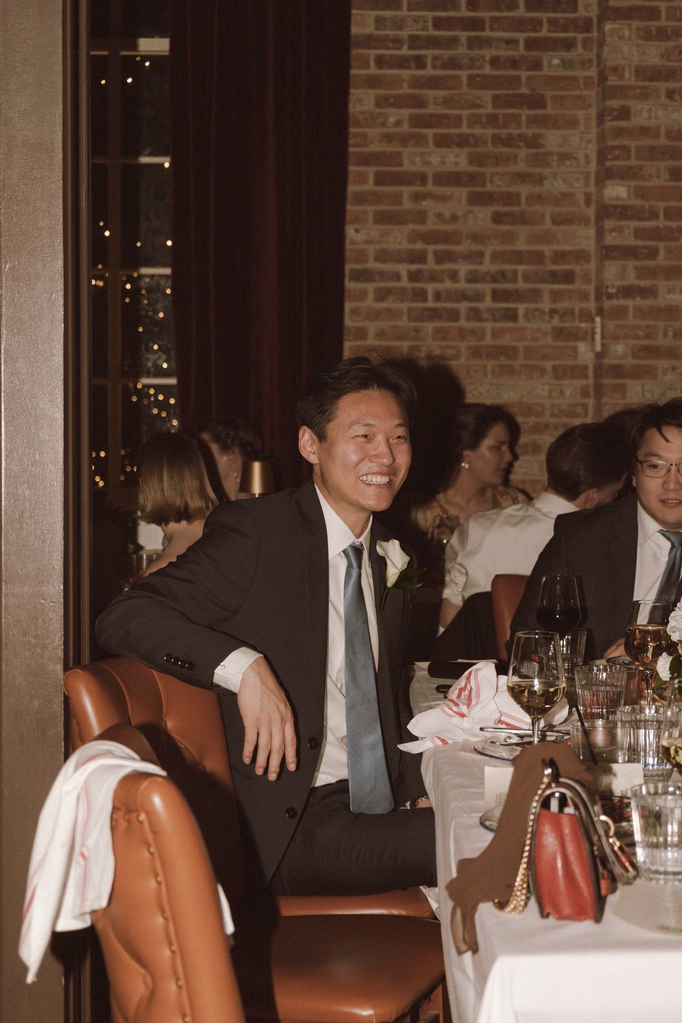 Smiling man in dark suit and light blue tie seated at elegant wedding reception dinner table with brick wall backdrop.