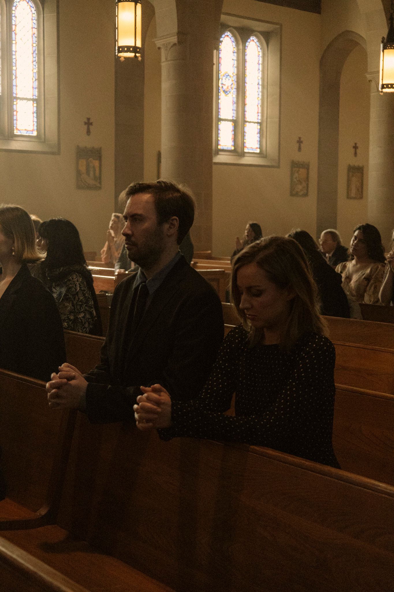 Man and woman with clasped hands pray solemnly in church pews during a wedding ceremony.