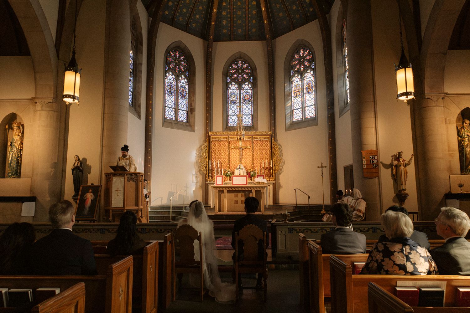 Wedding guests seated in wooden pews during ceremony in beautiful church with stained glass windows.