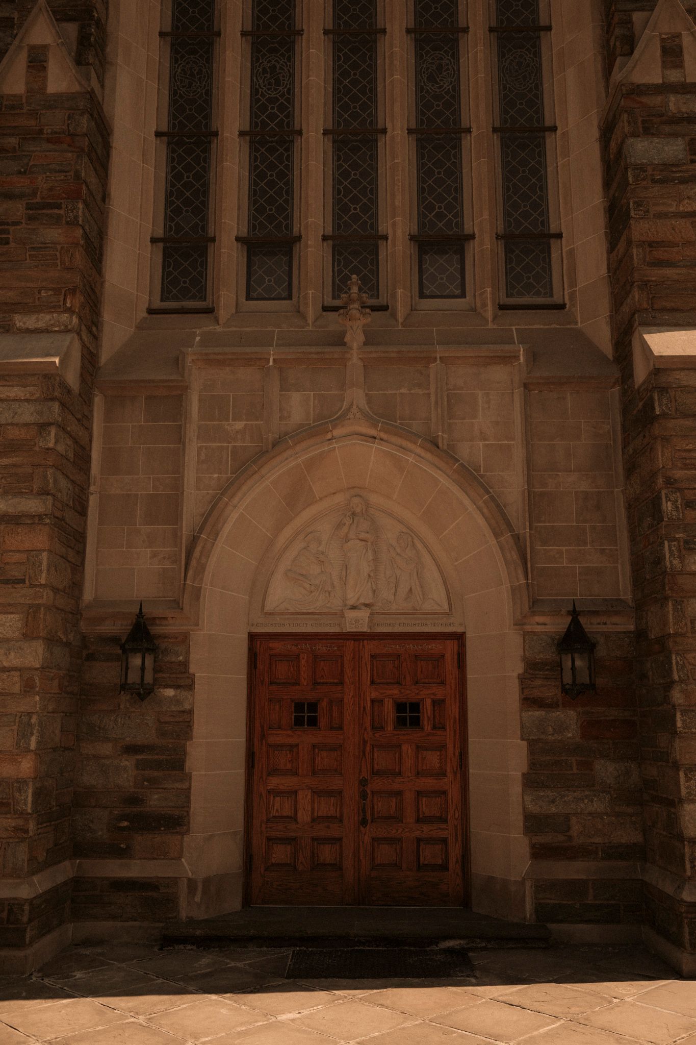 Gothic stone church entrance with ornate wooden double doors, arched relief sculpture, lantern sconces, and leaded glass windows above.