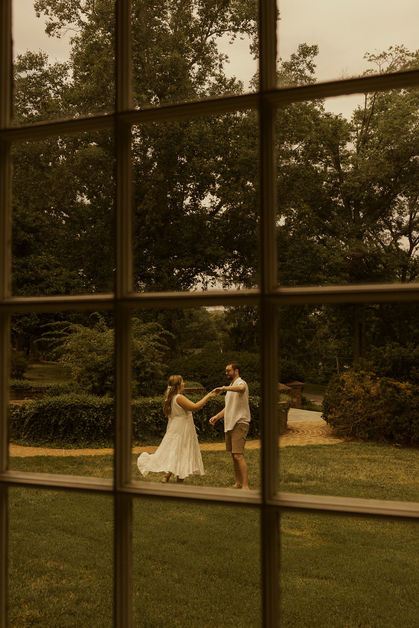 A couple dances in a lush garden, viewed through a large window, the woman in a white dress.