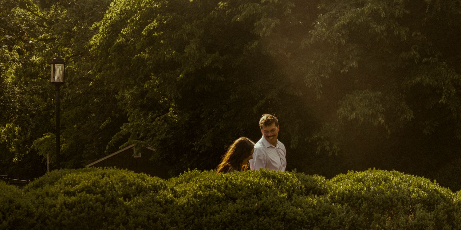 Two people sitting among lush green hedges in a sunlit garden surrounded by tall trees.
