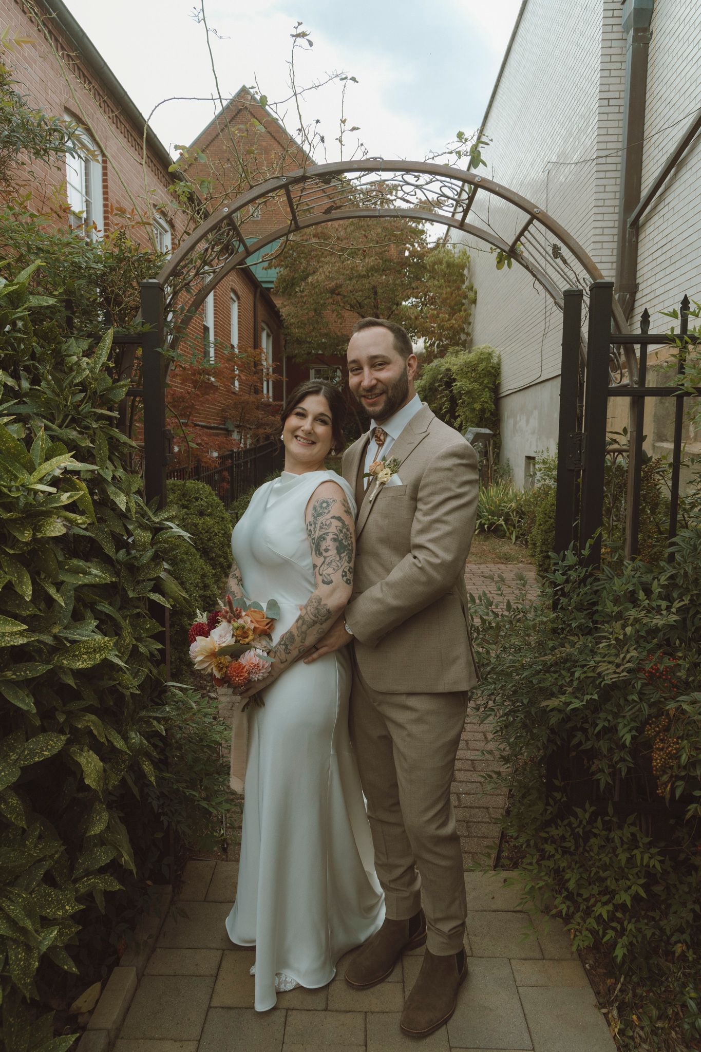 A wedding couple poses together in a garden pathway with brick buildings and greenery in the background.