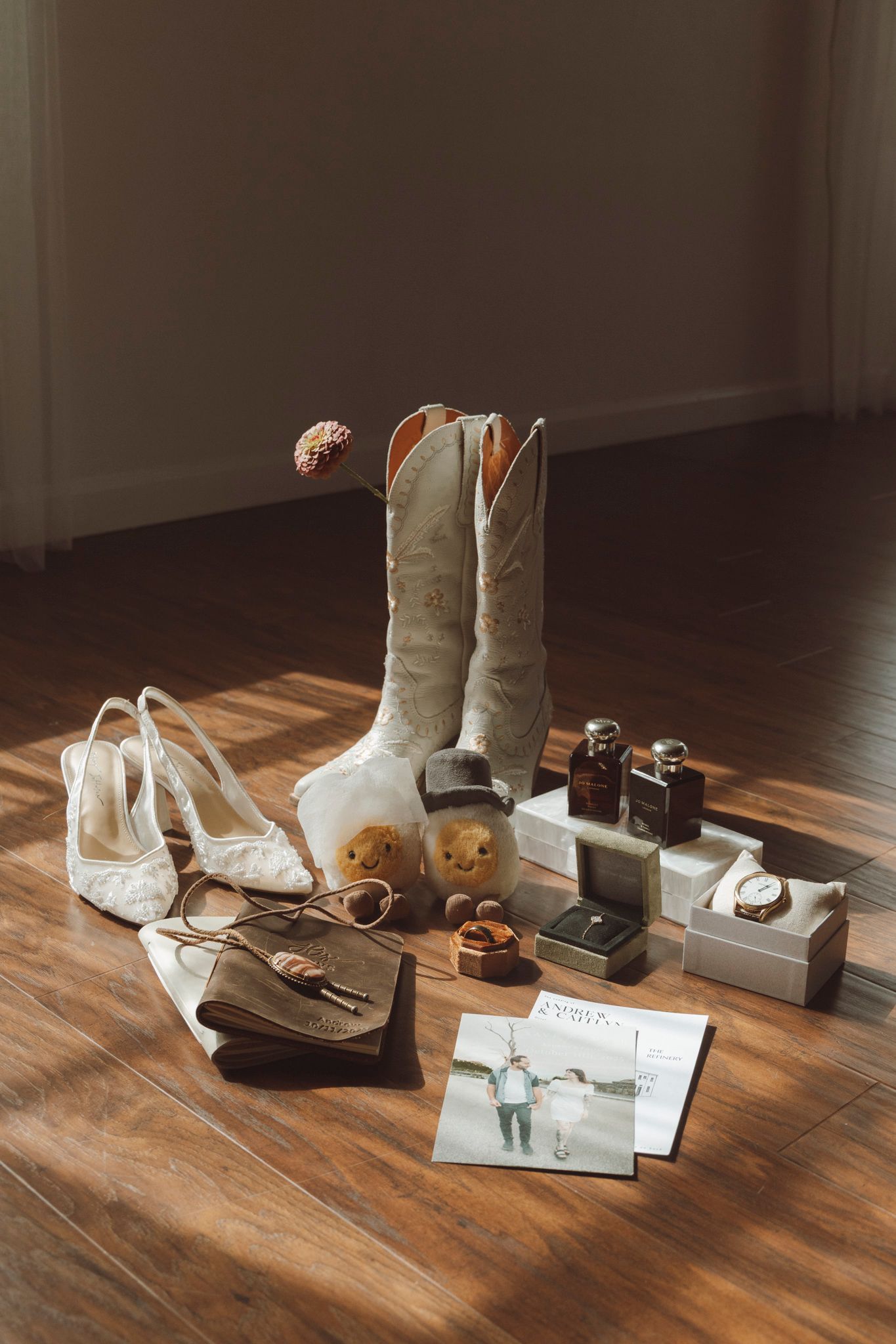 Wedding accessories including cowboy boots, heels, jewelry boxes, and invitations arranged on a wooden floor.