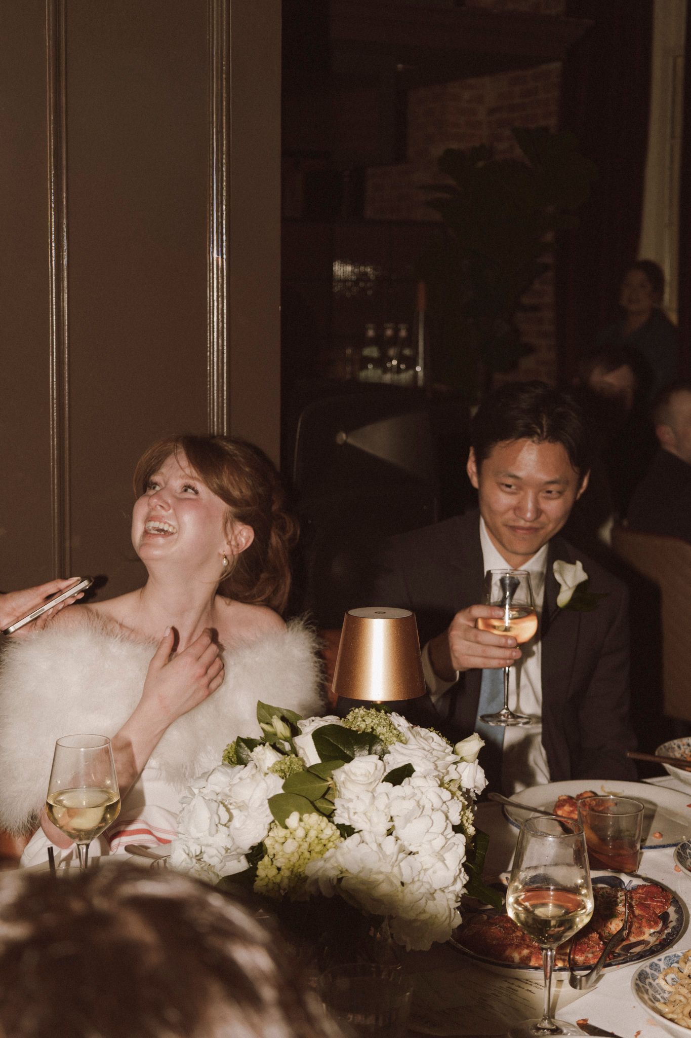 Bride and groom laughing and toasting with wine glasses at elegant wedding reception dinner table with white floral centerpiece.