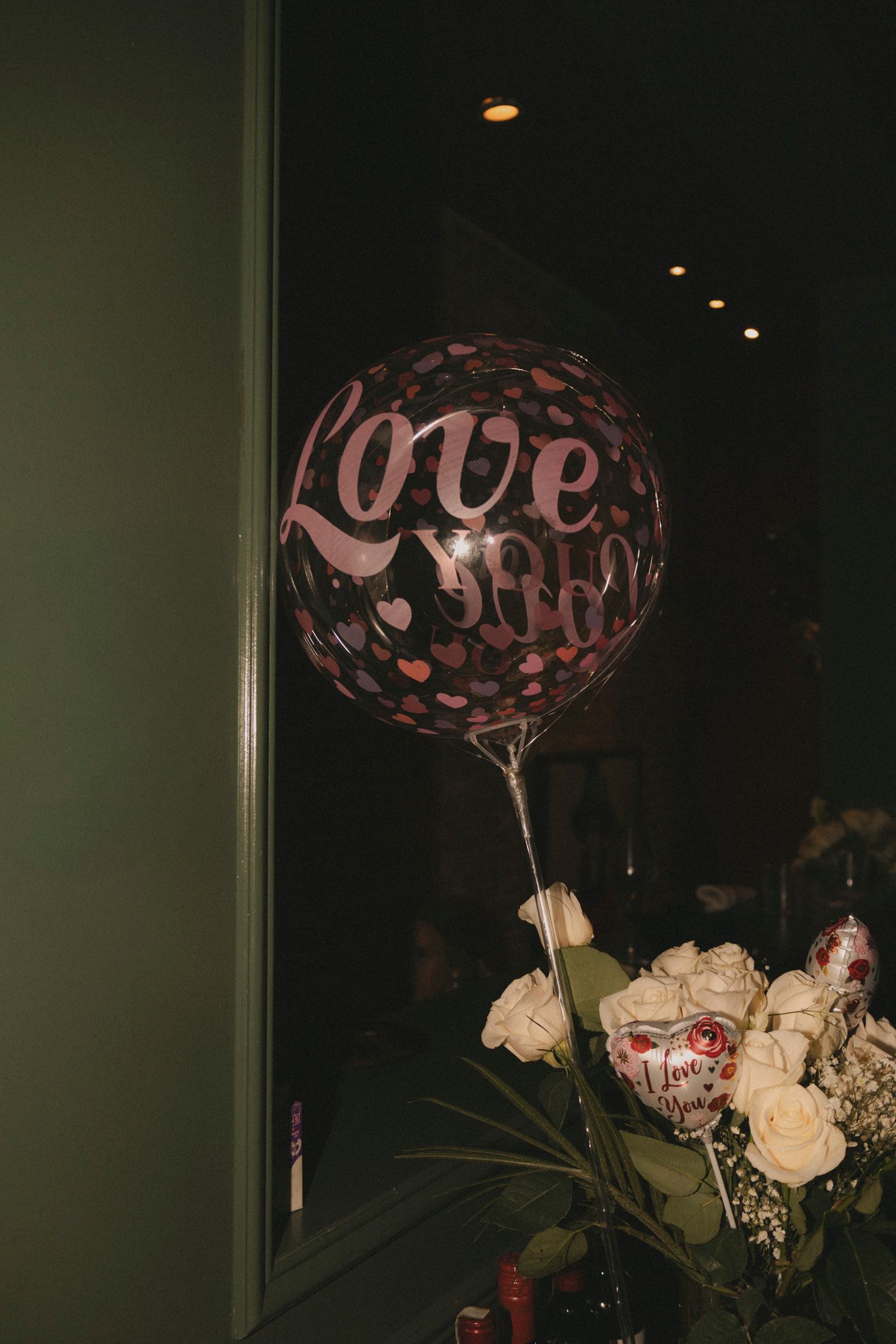 A large round Love You balloon with heart patterns beside white roses in a dimly lit romantic setting.