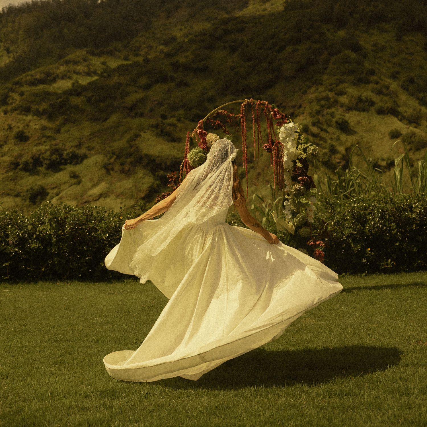 Bride in flowing white gown and veil twirling outdoors near floral arch with green hills backdrop.