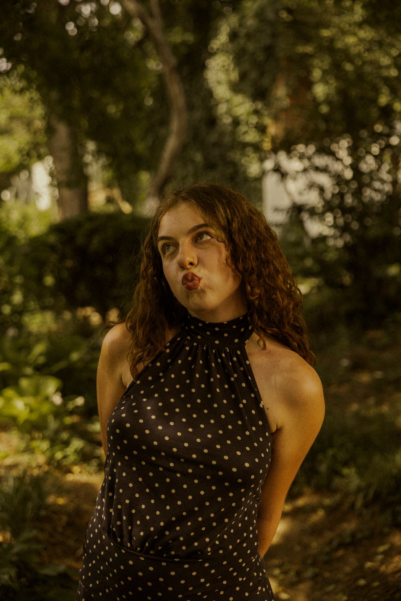 Young woman in black polka dot halter dress making a pouty face outdoors in a lush green garden setting.