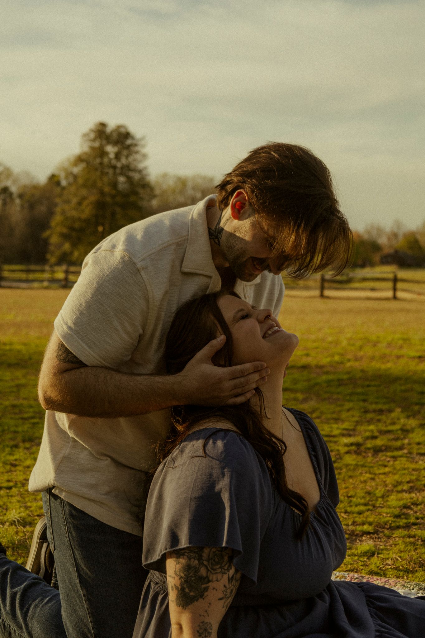 A couple shares a tender romantic moment outdoors at golden hour, man leaning down to kiss woman lying on grass.