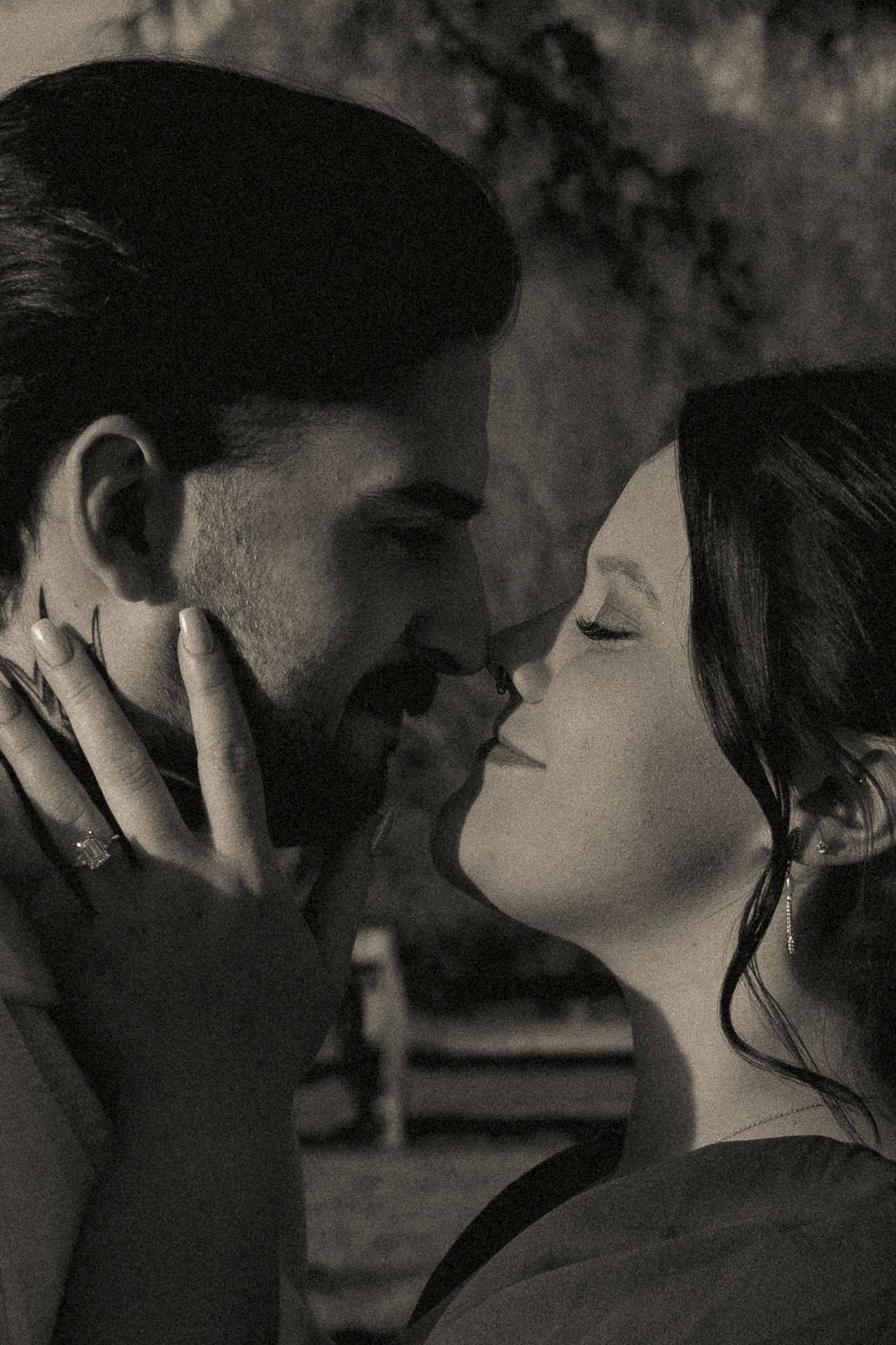 Black and white romantic photo of a couple almost kissing, woman gently holding mans face with engagement ring visible.