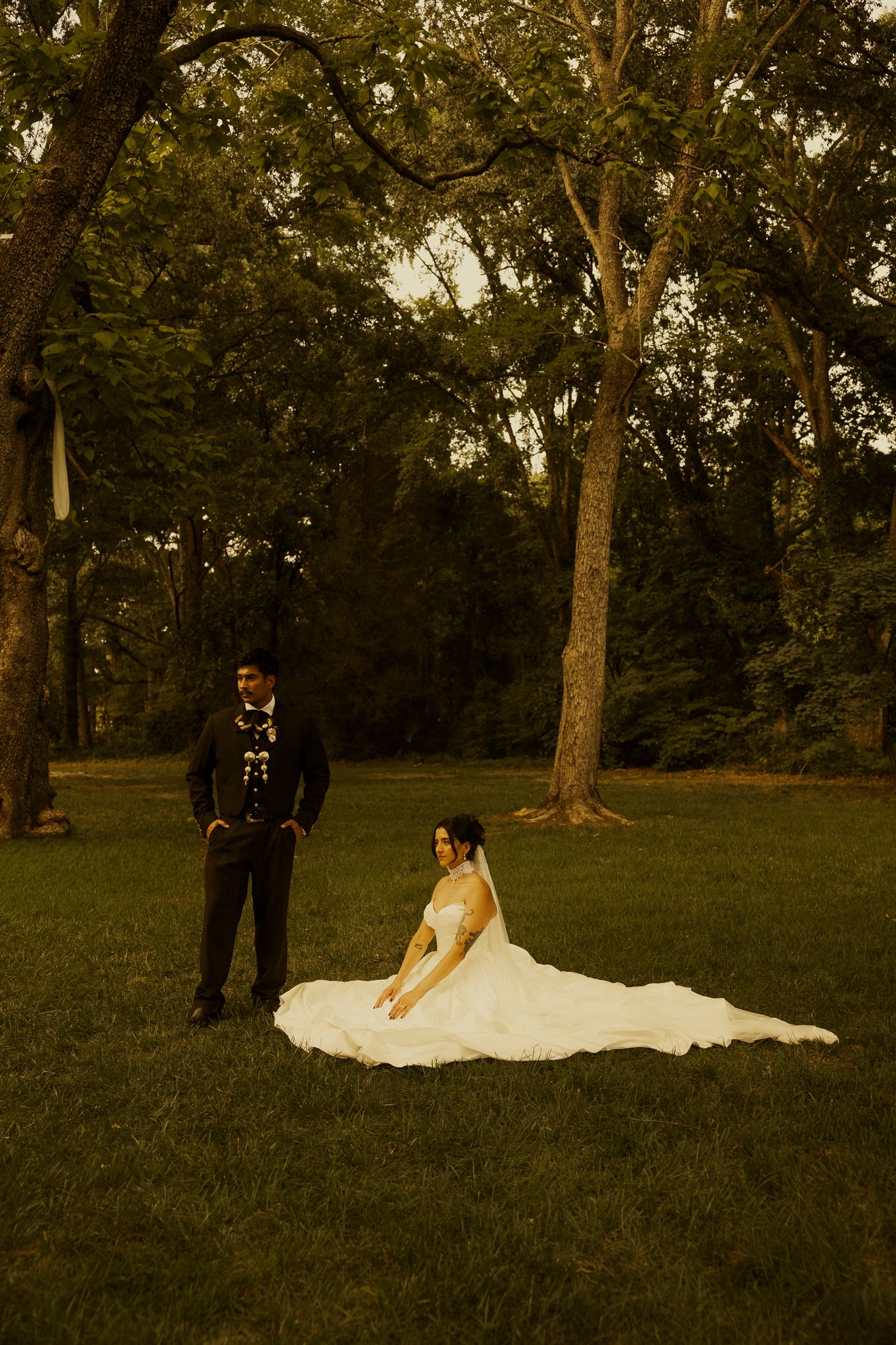 Bride in white gown sits on grass while groom stands nearby in lush green park during golden hour wedding portrait.