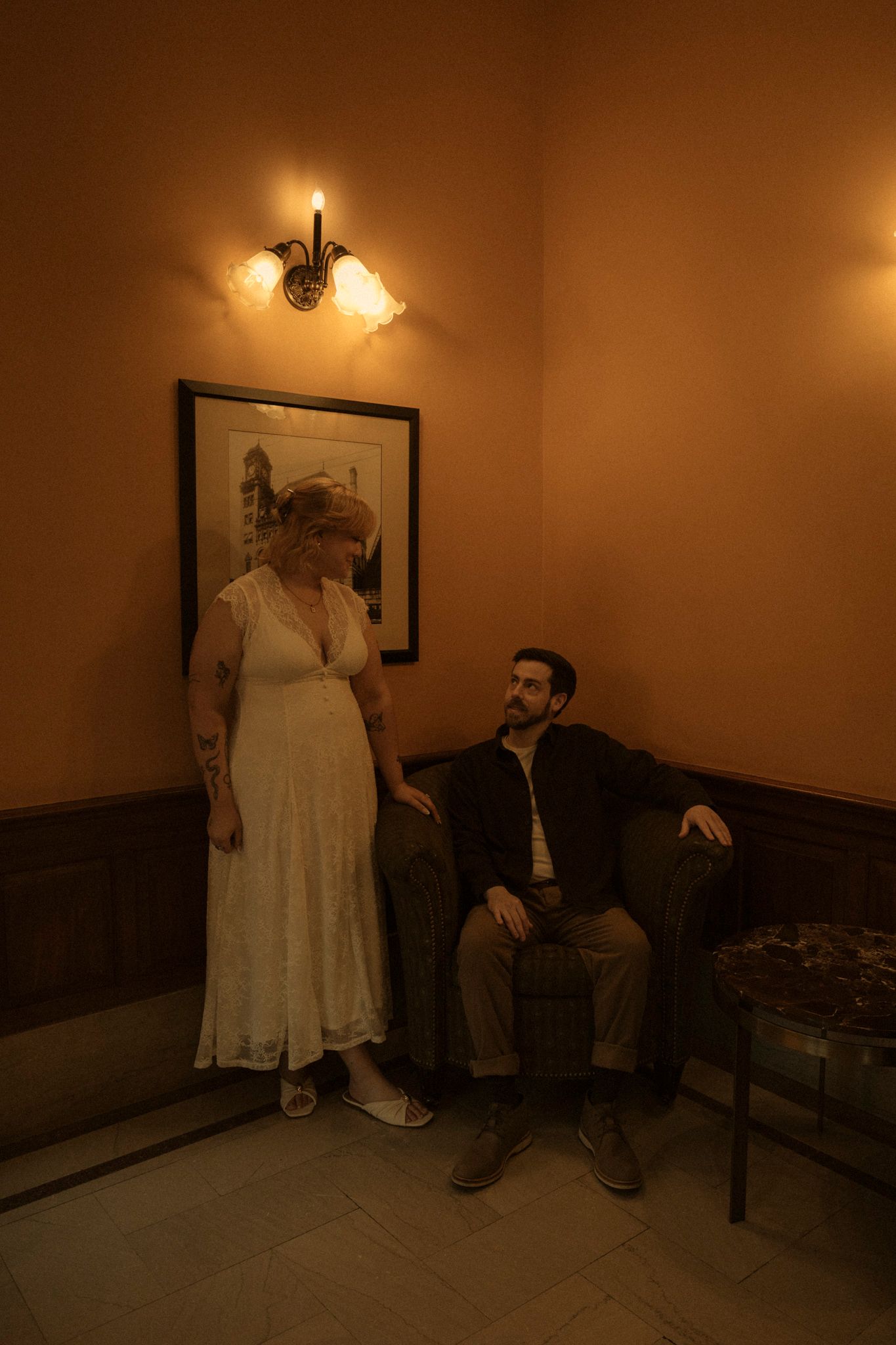 A figure in a white dress stands next to someone seated in a dimly lit hallway with vintage-style wall sconces.