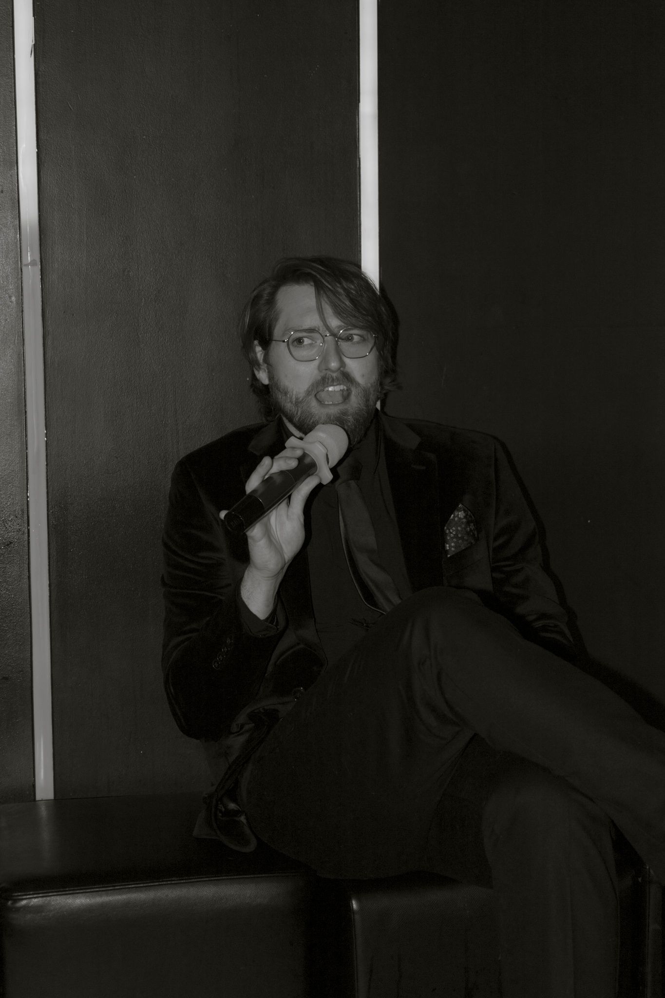 Black and white portrait of a performer in a dark suit sitting and singing with an expressive gesture.